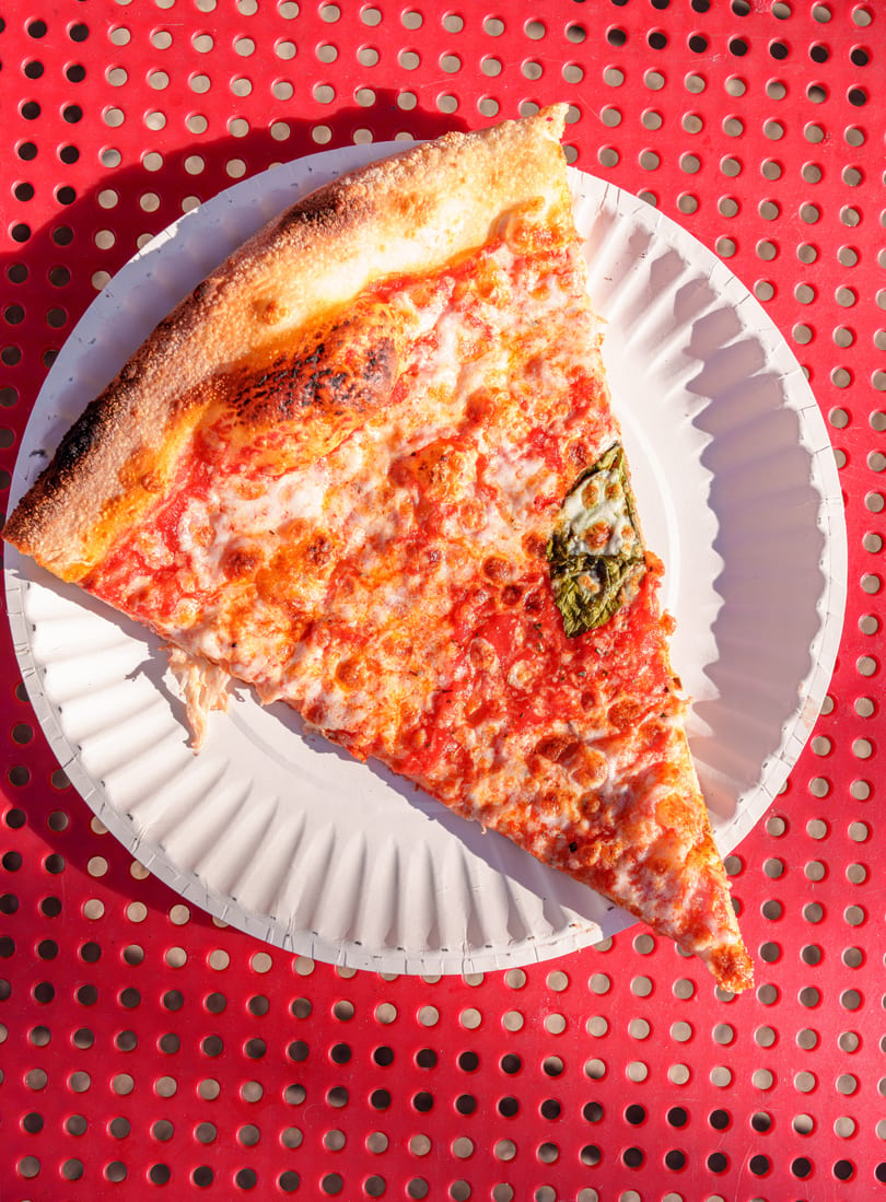 A single slice of cheese pizza with basil on a white paper plate, on a red perforated table.