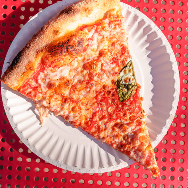 A single slice of cheese pizza with basil on a white paper plate, on a red perforated table.