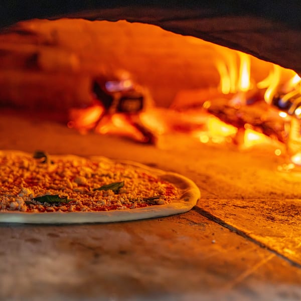 Uncooked pizza with cheese and basil leaves baking inside a fiery wood-fired oven.