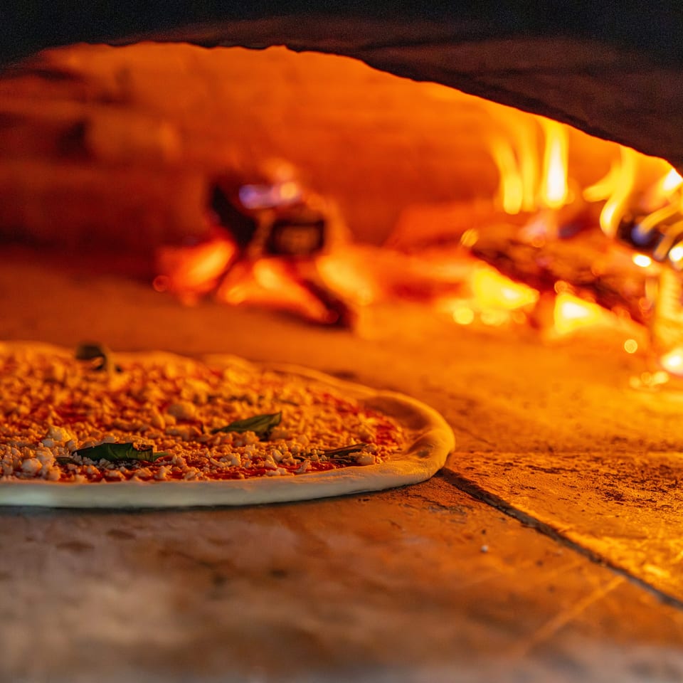 Uncooked pizza with cheese and basil leaves baking inside a fiery wood-fired oven.