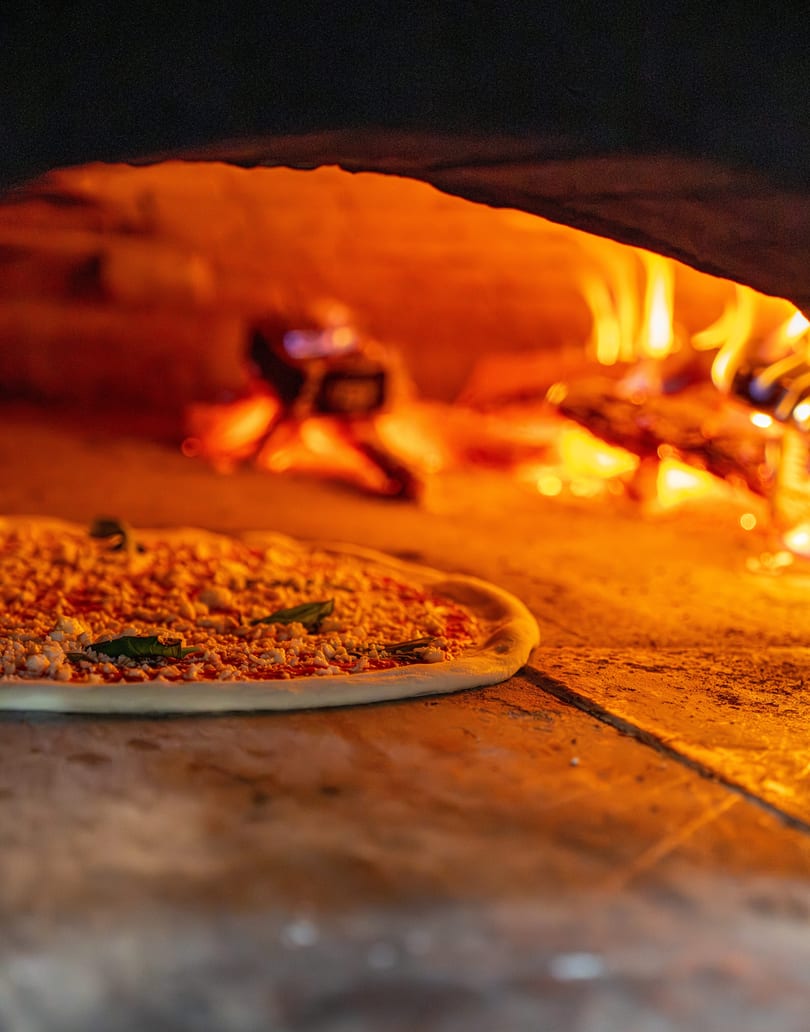 Uncooked pizza with cheese and basil leaves baking inside a fiery wood-fired oven.