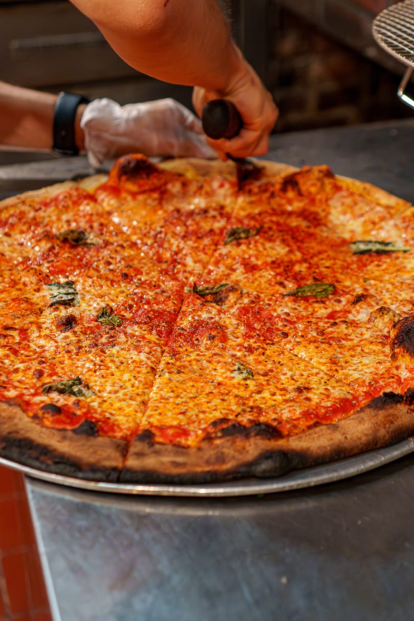 A chef's hands slice a large, cheesy pizza with basil on a metal tray.