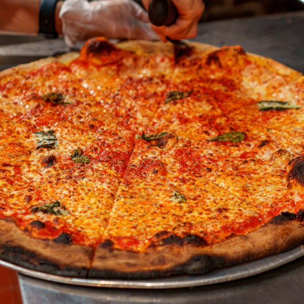 A chef's gloved hand uses a pizza cutter to slice a large, cheesy Margherita pizza.
