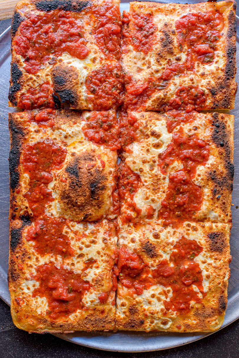 A square Detroit-style pizza with crispy charred edges, topped with melted cheese and dollops of red sauce.