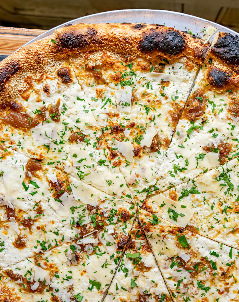 Close-up of a white pizza with caramelized onions, parmesan, and parsley, sliced on a metal tray.