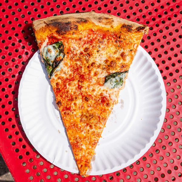 A large slice of cheese and basil pizza on a white paper plate, on a red perforated table.