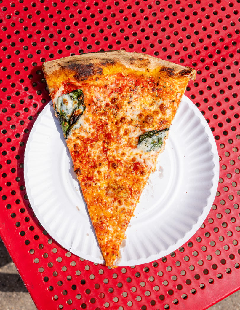 A large slice of cheese and basil pizza on a white paper plate, on a red perforated table.