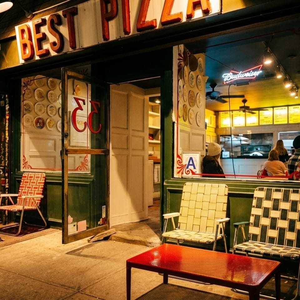 Exterior of a pizza shop at night, with outdoor seating and customers visible inside.