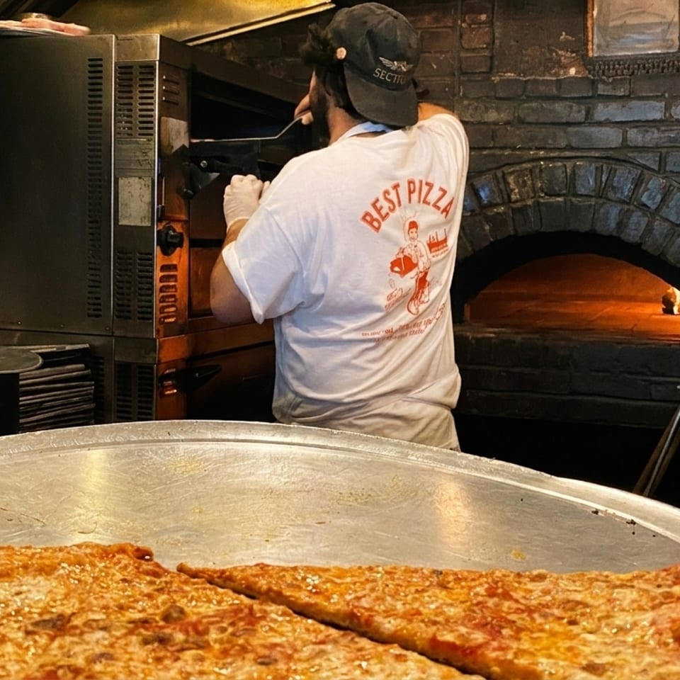 A pizza chef in a "Best Pizza" shirt works near a brick oven, with a large pizza in the foreground.