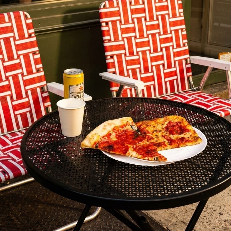 Two slices of pizza, a drink, and a beer can on a black patio table with red and white chairs.