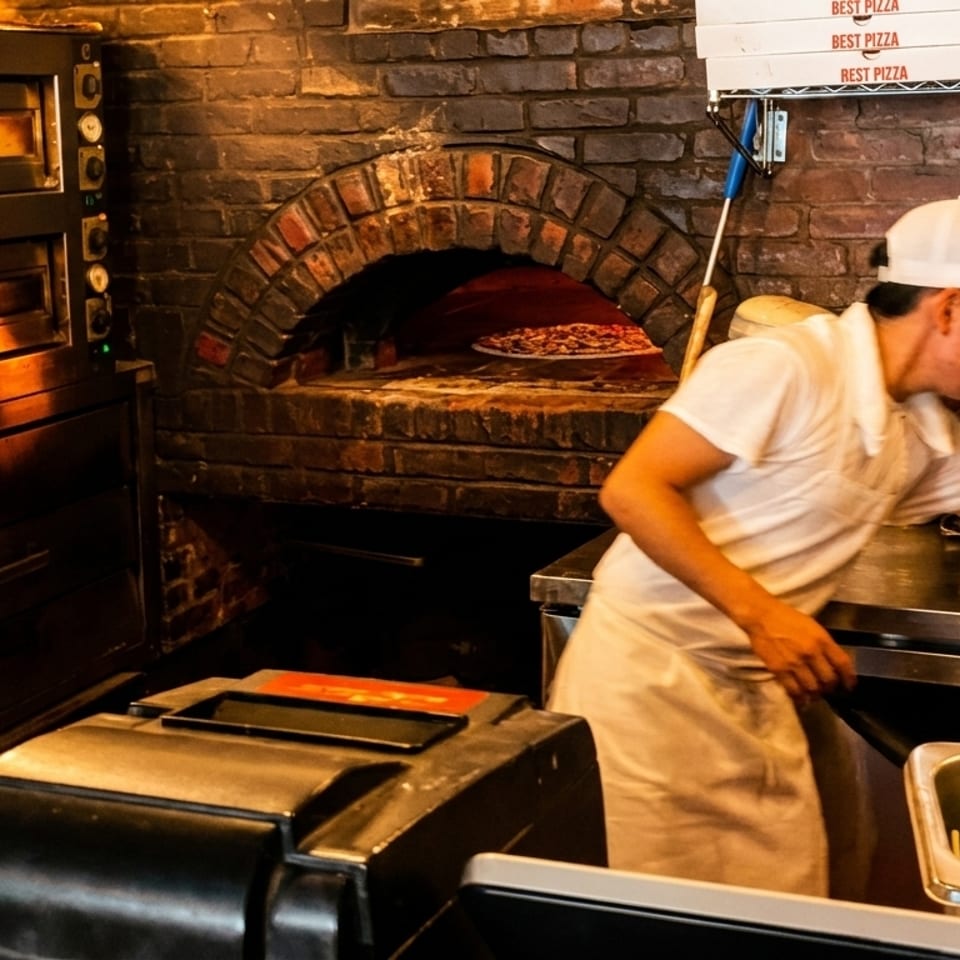 A pizza chef prepares a pizza next to a brick oven and stainless steel equipment.