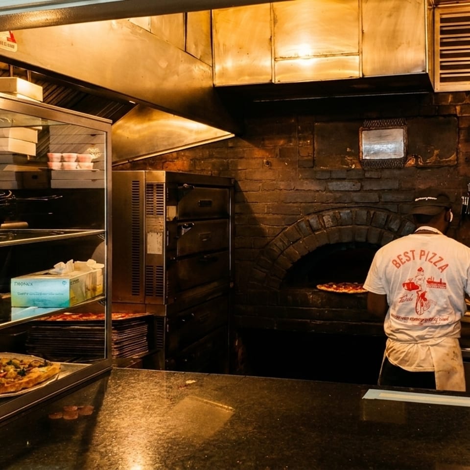 A chef in a "Best Pizza" shirt puts a pizza into a brick oven in a restaurant kitchen.