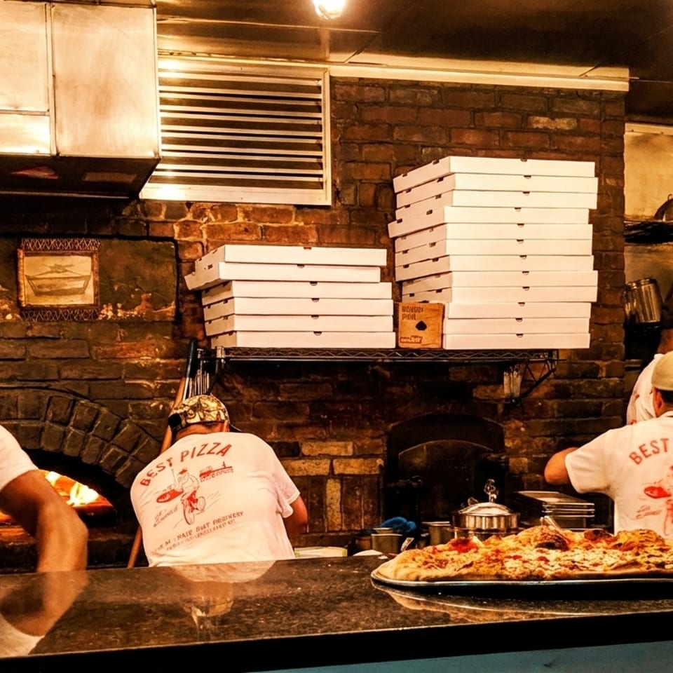 Pizzaiolos at Best Pizza working in a brick oven kitchen with pizzas and stacked boxes.